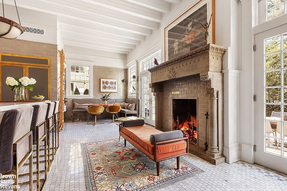 This interior shot showcases a stylish living room with a prominent fireplace, a daybed, and a dining area. The room features white tiled floors, exposed ceiling beams, and large windows that provide ample natural light. The overall aesthetic is elegant and inviting, blending classic architectural details with modern furnishings.
