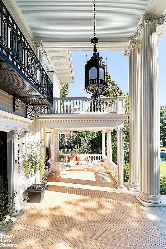 This image showcases a grand, covered patio or balcony area of a stately home. The space features herringbone brick flooring, substantial white columns with ornate capitals, and a hanging lantern. Comfortable outdoor seating is visible in the background, suggesting a relaxing and inviting atmosphere.