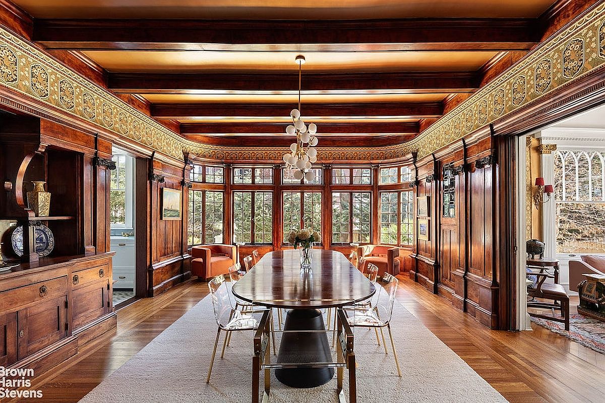 This is a dining room featuring rich wood paneling, a coffered ceiling with decorative trim, and a large bay window offering natural light. A long, polished dining table is centered in the room, surrounded by modern chairs, and a light-colored rug anchors the space. The room exudes a sense of classic elegance and sophistication.