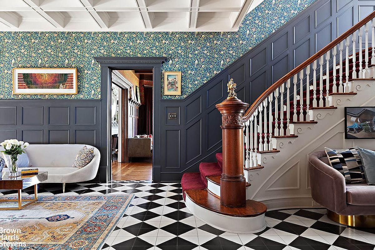 This interior shot showcases a grand hallway with a striking black and white checkered floor, complemented by a patterned rug. A staircase with a red runner and white balusters ascends to the upper level, while elegant furnishings such as a curved sofa and a side table add to the sophisticated ambiance. The walls feature dark wood paneling and floral wallpaper, creating a luxurious and inviting entryway.