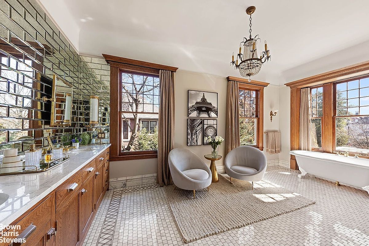 This is a luxurious primary bathroom featuring a double vanity with a mirrored backsplash, a freestanding bathtub, and two armchairs with a small table between them. The room is well-lit by natural light from the windows, which are framed by wooden trim and curtains, and an ornate chandelier hangs from the ceiling. The overall impression is elegant and spacious.