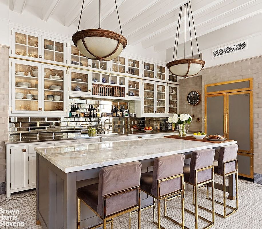 This is a well-lit kitchen featuring a large island with marble countertops and seating for four. The kitchen includes custom cabinetry with glass fronts, mirrored backsplash, and high-end appliances, creating a luxurious and functional space. Two pendant lights hang above the island, adding to the room's elegance.