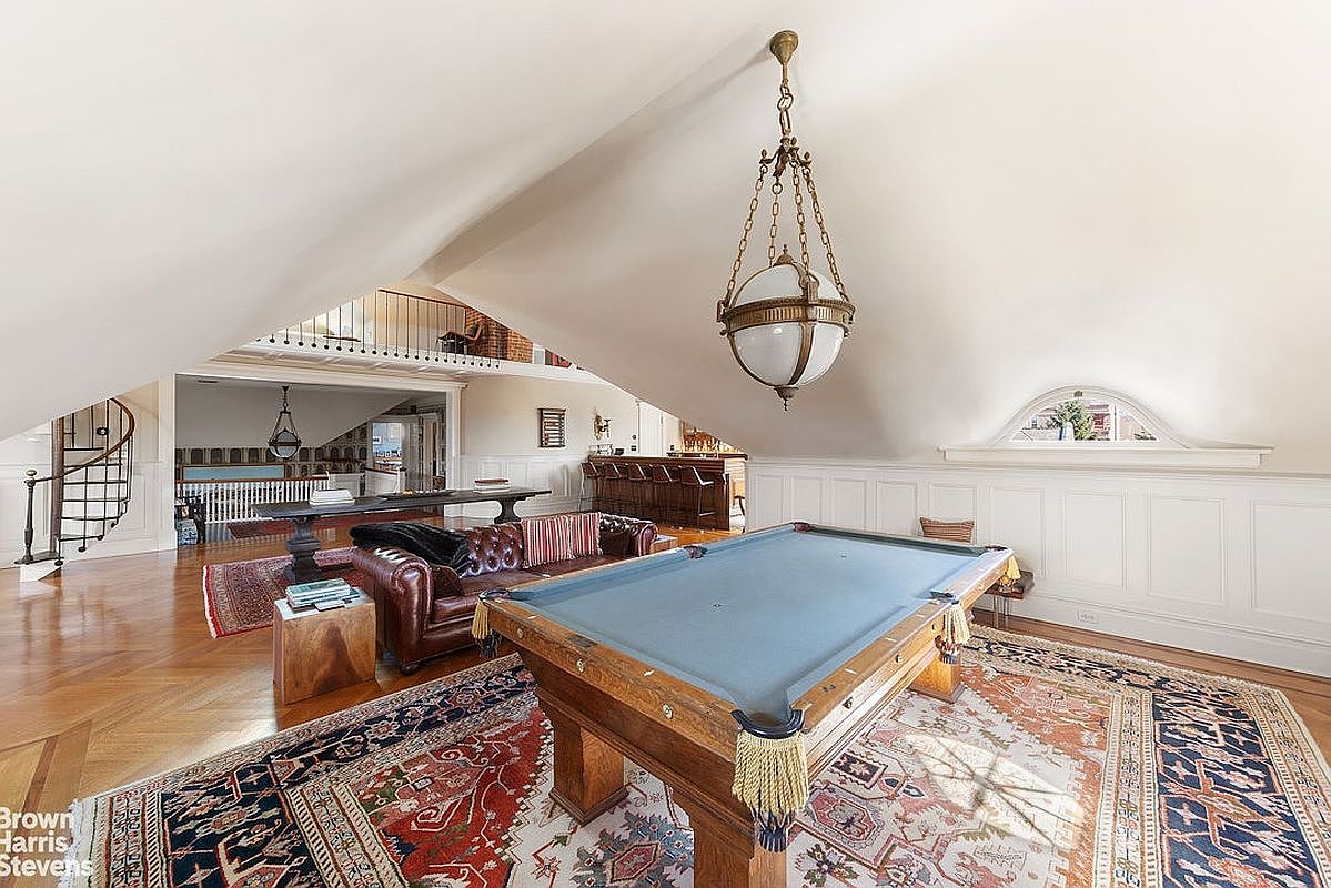 This is a spacious living room featuring a pool table as a central element, complemented by a leather sofa and a wooden side table. The room is illuminated by a vintage-style chandelier, and the hardwood floors are adorned with ornate rugs. The space also includes a bar area and a staircase leading to an upper level, creating a luxurious and inviting atmosphere.