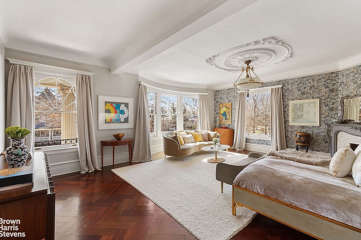 This primary bedroom features a blend of classic and contemporary design elements. The room boasts a curved bay window area with a comfortable sofa, a decorative ceiling medallion with a chandelier, and a bed with a neutral-toned comforter. The walls are adorned with patterned wallpaper on one side and neutral paint on the other, creating a sophisticated and inviting atmosphere.