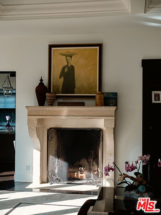 This interior view highlights a classic stone fireplace mantel serving as the focal point of the room, adorned with a framed portrait painting and decorative vases. To the right, a vibrant orchid arrangement adds a touch of color, while the adjacent doorway leads to a glimpse of a kitchen area. The composition is captured from a slightly elevated perspective, emphasizing the elegant, neutral-toned architectural details and the sophisticated atmosphere of the living space.