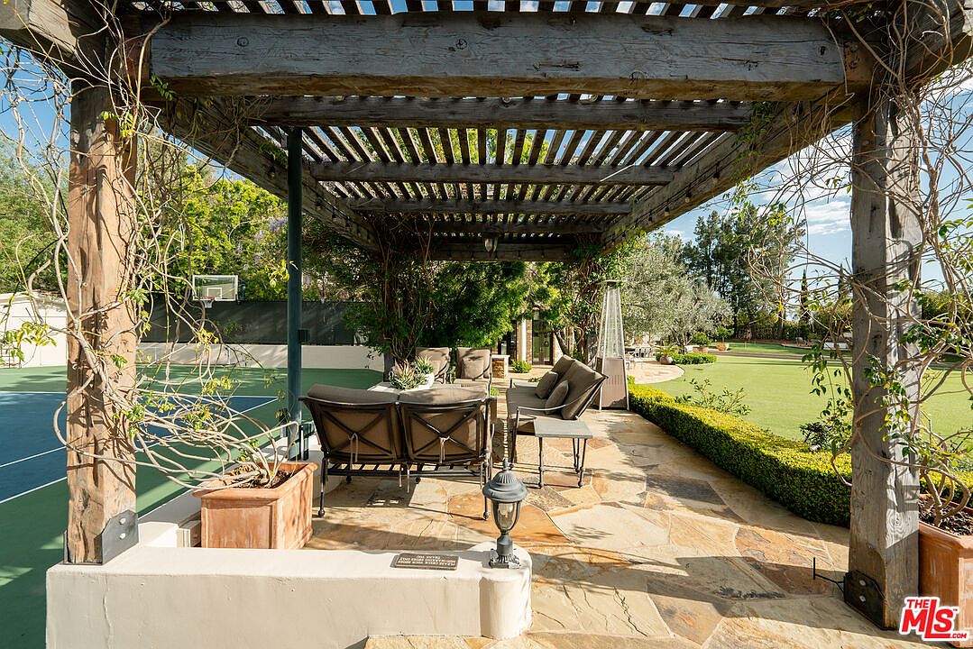 This outdoor living space features a rustic wooden pergola providing shade over a comfortable seating area with cushioned furniture. The patio is paved with natural stone and overlooks a well-manicured lawn and a private tennis court. The setting offers a serene and luxurious atmosphere, perfect for relaxation or entertaining guests in a resort-like backyard environment.