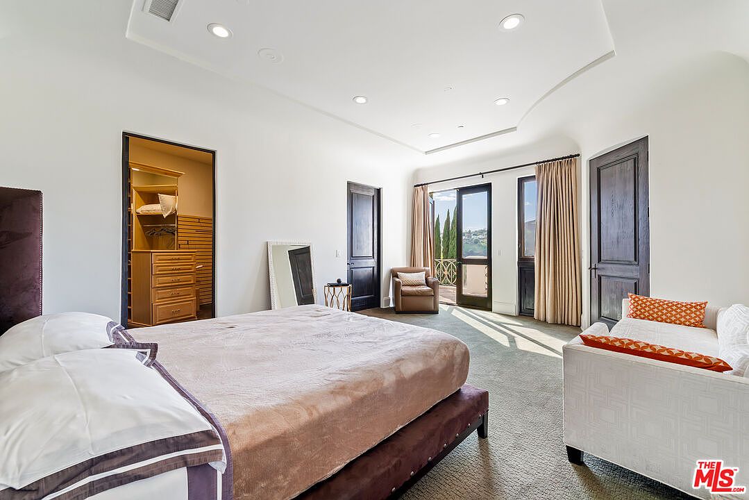 This spacious primary bedroom features a neutral color palette, plush carpeting, and a large bed centered in the room. To the left, an open doorway reveals a walk-in closet, while a seating area with a white sofa and a leather armchair sits near the large glass doors that open to a balcony. The room is illuminated by recessed lighting and natural light, creating an airy and sophisticated atmosphere.