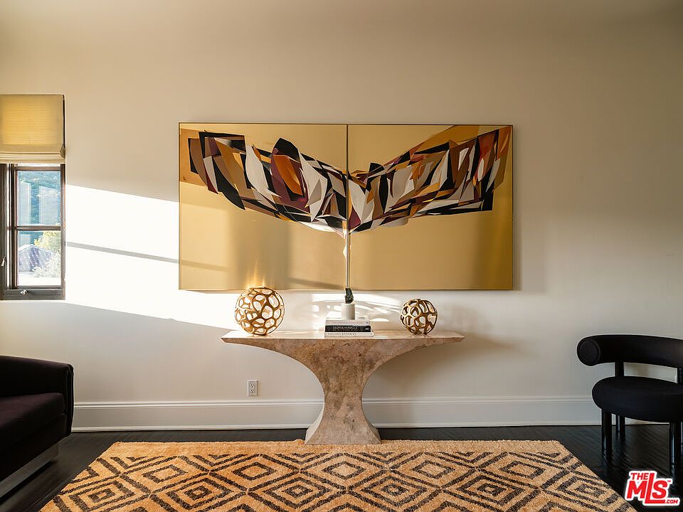 This interior space features a striking, large-scale abstract art piece mounted above a sculptural, light-toned stone console table. Two decorative, spherical wire sculptures sit on the console, which is positioned centrally against a neutral wall. A patterned area rug anchors the space, while a dark armchair is visible on the right, creating a sophisticated and curated aesthetic.