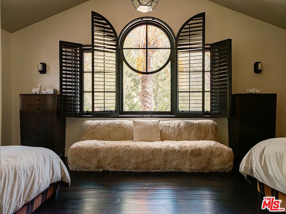 This bedroom features a central, plush, textured sofa positioned beneath a large, arched window with dark wooden shutters. The room is framed by two beds on either side, dark wood flooring, and symmetrical wall sconces, creating a cozy and balanced atmosphere. The perspective is centered, highlighting the architectural window as the focal point of the space.