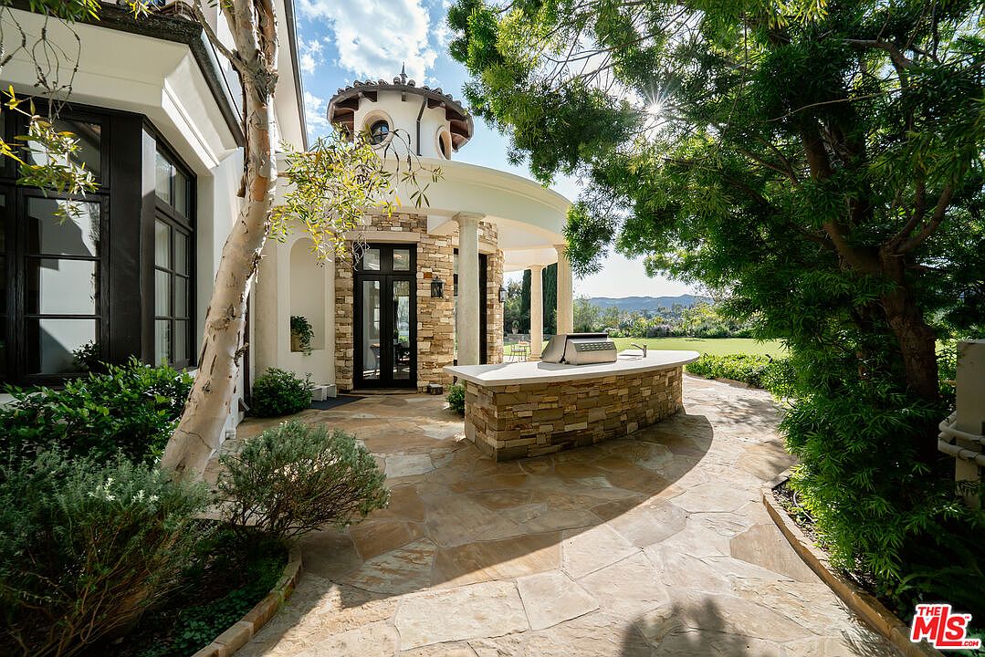 This outdoor living space features a curved stone-clad bar with a built-in grill, set upon a flagstone patio. The area is framed by a Mediterranean-style home with a distinctive rotunda, tall columns, and dark-framed glass doors. Lush trees and greenery surround the patio, creating a serene and private atmosphere perfect for outdoor entertaining.
