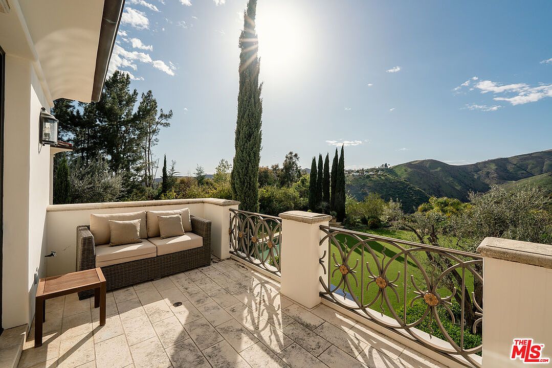 This elevated balcony offers a serene outdoor living space with a wicker sofa and a small side table set against a backdrop of rolling hills and lush greenery. The space features light-toned stone flooring and an ornate metal railing that provides an unobstructed view of the landscape below. Tall, slender cypress trees frame the horizon, enhancing the Mediterranean-inspired aesthetic of this peaceful retreat.