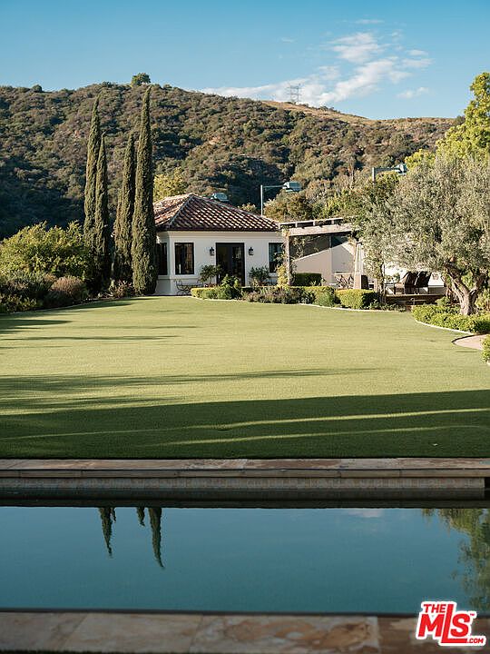 This serene outdoor scene features a calm swimming pool in the foreground, reflecting the tall, slender cypress trees and the clear blue sky. Beyond the pool, a manicured lawn leads to a charming white structure with a tiled roof, set against a backdrop of rolling, sun-drenched hills. The composition offers a peaceful, resort-like atmosphere, highlighting the seamless integration between the landscaped grounds and the natural hillside terrain.