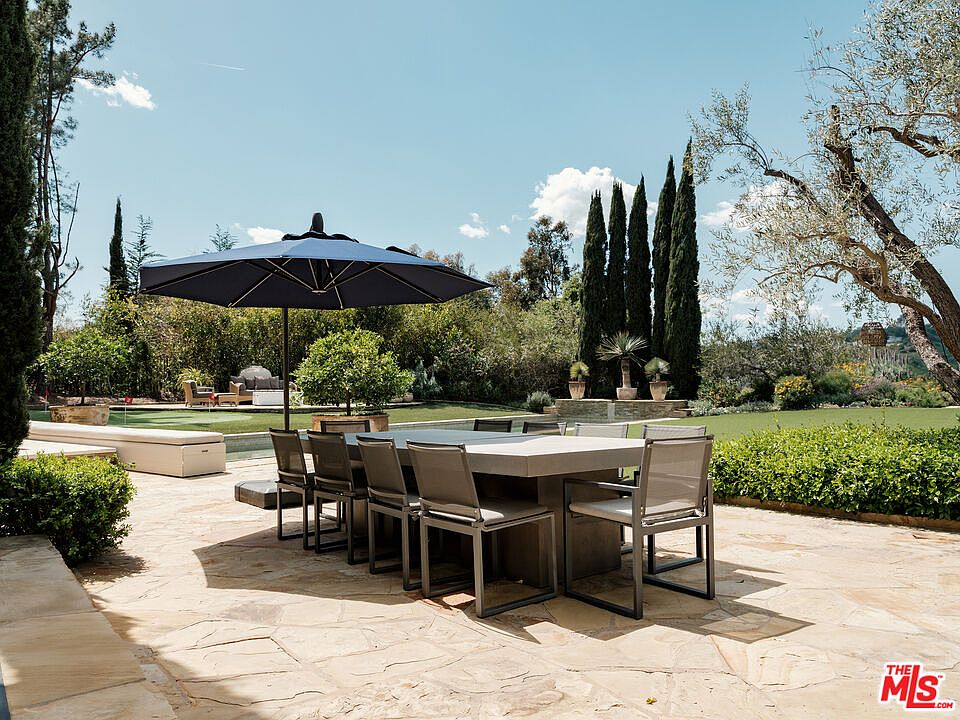 This expansive outdoor patio features a large, modern dining table set with eight chairs, shaded by a prominent dark blue umbrella. The stone-paved terrace overlooks a manicured lawn, mature landscaping, and tall cypress trees, creating a serene and private atmosphere. The scene is captured from a ground-level perspective, emphasizing the seamless transition between the dining area and the lush backyard environment.