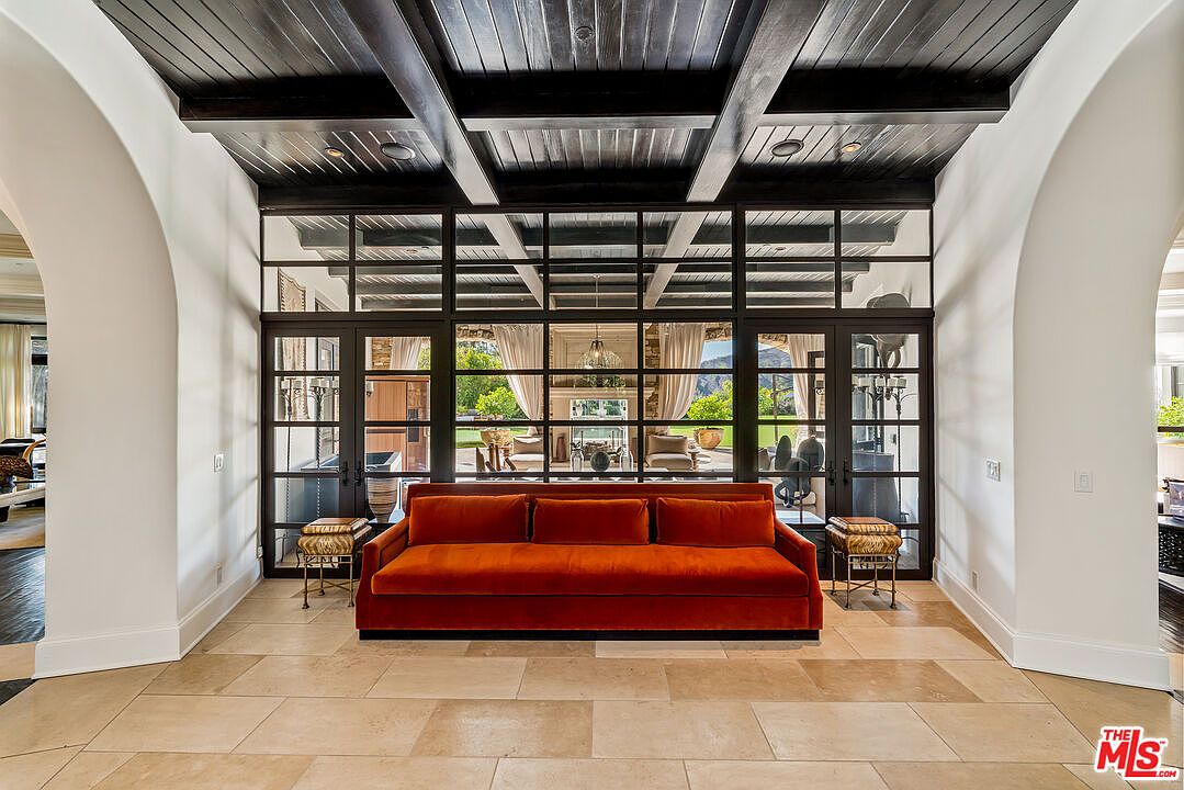 This elegant living area features a vibrant orange velvet sofa centered against a striking wall of black-framed glass panels that offer a view into an adjacent room. The space is defined by a dark, coffered wood ceiling and light-toned stone flooring, creating a sophisticated contrast. The symmetrical layout and arched doorways on either side contribute to a sense of architectural balance and refined luxury.