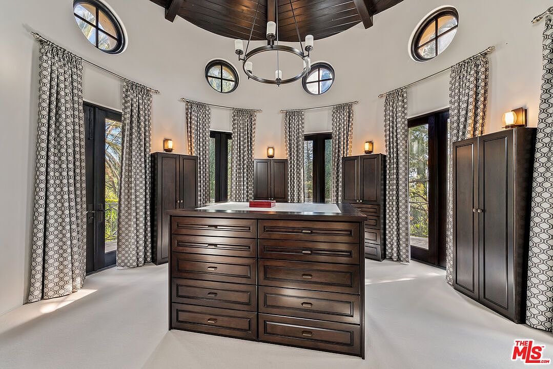 This spacious, circular walk-in closet features a large, dark wood central island with numerous drawers for storage. The room is accented by a dark wood coffered ceiling, a central chandelier, and multiple tall windows that provide natural light. The space is organized with dark wood cabinetry along the walls and is finished with neutral carpeting, creating a sophisticated and functional dressing area.