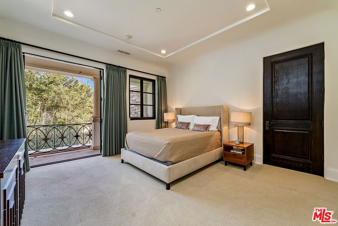 This spacious primary bedroom features a neutral color palette, plush carpeting, and a tray ceiling with recessed lighting. A large glass door opens onto a private balcony with a decorative metal railing, providing a scenic view of the surrounding trees. The room is furnished with a bed centered against the wall, flanked by two wooden nightstands with matching lamps, and features a dark wood door that adds a touch of contrast to the light-toned walls.