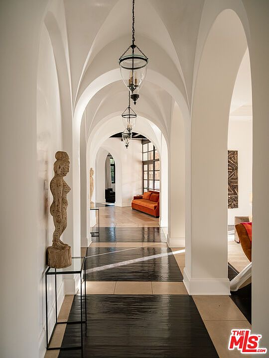 This elegant hallway features a series of dramatic, white-painted arched openings that create a sense of depth and architectural grandeur. The floor is finished with a combination of light-toned stone tiles and a dark, reflective inlay that draws the eye toward the living space at the end of the corridor. A tall, carved wooden sculpture stands on a minimalist glass pedestal in the foreground, while classic lantern-style pendant lights hang from the vaulted ceiling.