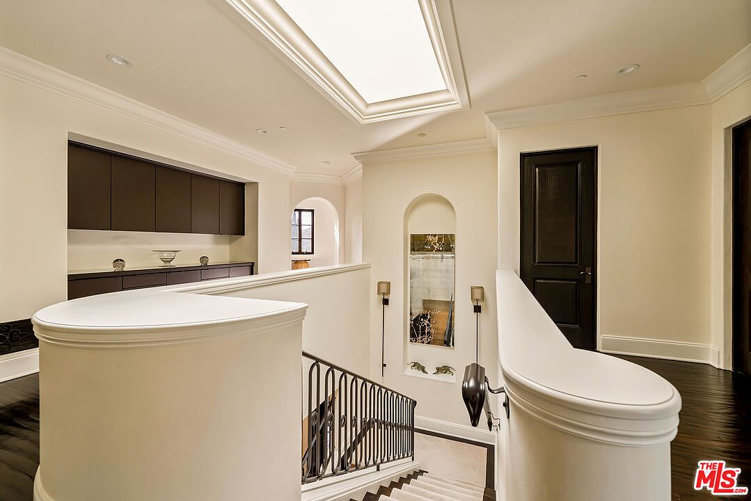 This elegant upper-level landing features a curved white staircase railing and dark wood flooring, creating a sophisticated transition between levels. The space is illuminated by a large rectangular skylight and includes a built-in dark cabinetry unit for storage or display. A decorative arched wall niche with a mirror adds architectural interest, while a dark door provides a sharp contrast to the neutral walls.