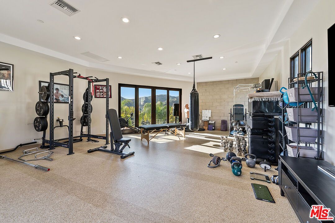 This spacious home gym features a comprehensive setup including a power rack, a punching bag, a massage table, and various free weights. The room is illuminated by recessed lighting and features large glass doors that provide a clear view of the surrounding hills. The neutral color palette and durable flooring create a functional and professional workout environment.