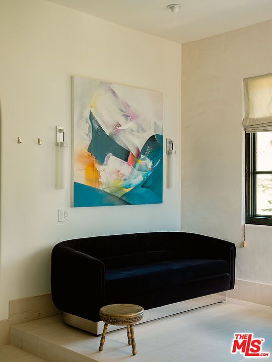 This elegant seating area features a plush, dark velvet sofa with a metallic base, positioned against a crisp white wall adorned with a large, vibrant abstract painting. Flanking the artwork are two modern, vertical wall sconces, while a small, ornate brass-toned stool sits in the foreground. The space is set on a raised platform, creating a distinct lounge area that feels both sophisticated and intimate.