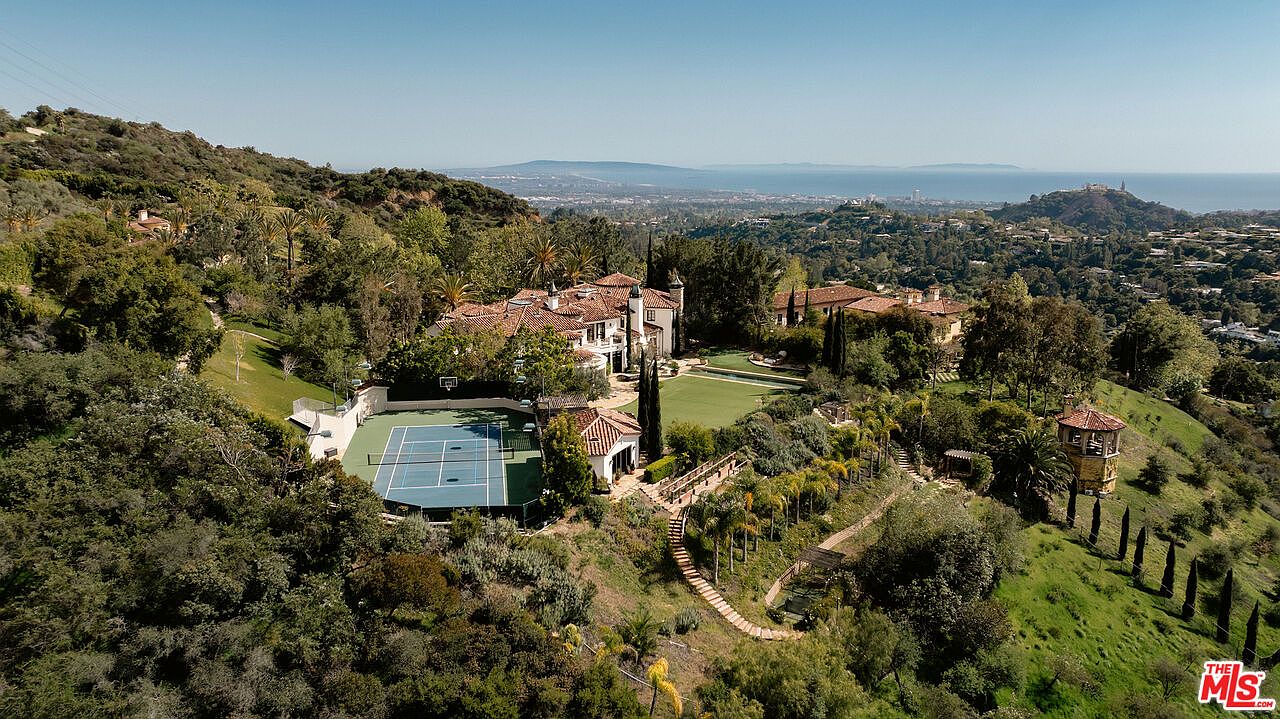 This expansive aerial view captures a sprawling Mediterranean-style estate nestled within a lush, hilly landscape. The property features a private tennis court, a manicured lawn, and a multi-level residence with a terracotta-tiled roof, all set against a backdrop of rolling hills and a distant coastal horizon. The perspective highlights the secluded, resort-like nature of the grounds and the integration of the home into its natural environment.