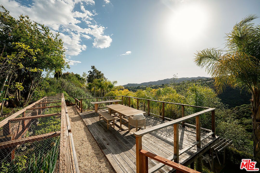 This elevated wooden deck offers a serene outdoor dining space with a long table and bench seating, overlooking a lush, hilly landscape. To the left, a wire-fenced garden area adds a rustic touch, while the open railing ensures unobstructed views of the surrounding greenery and distant mountains. The scene is bathed in bright, natural sunlight, creating an inviting atmosphere for relaxation or entertaining.