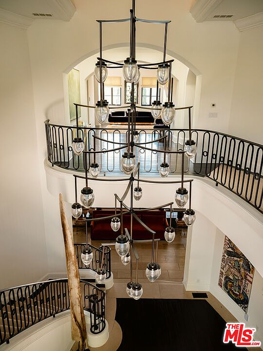 This high-angle perspective captures a grand foyer featuring a dramatic, multi-tiered chandelier with numerous glass pendants suspended over a curved staircase. The space showcases a classic architectural design with a wrought-iron railing that follows the sweeping curve of the landing, overlooking a lower level with a dark-toned sofa. The neutral color palette and open layout create an airy, sophisticated atmosphere that emphasizes the home's vertical scale and elegant transition between floors.