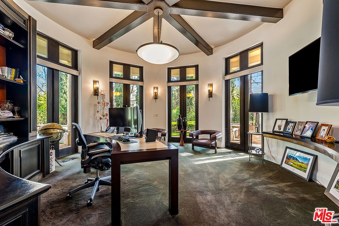 This sophisticated home office features a dark wood desk positioned centrally under a striking vaulted ceiling with exposed beams and a large pendant light. The room is surrounded by tall windows and glass doors that provide abundant natural light and views of the lush greenery outside. A built-in bookshelf, a leather armchair, and a console table displaying framed photos complete this professional yet comfortable workspace.