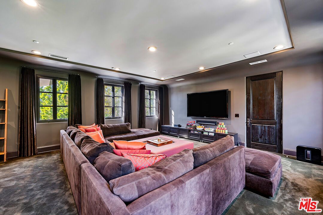This spacious media room features a large, plush sectional sofa arranged around a central ottoman, creating a comfortable and inviting atmosphere. The room is anchored by a wall-mounted television above a long media console, with dark walls and recessed lighting providing a sophisticated, theater-like ambiance. Three windows with dark curtains allow natural light to filter in, balancing the deep tones of the room's decor.
