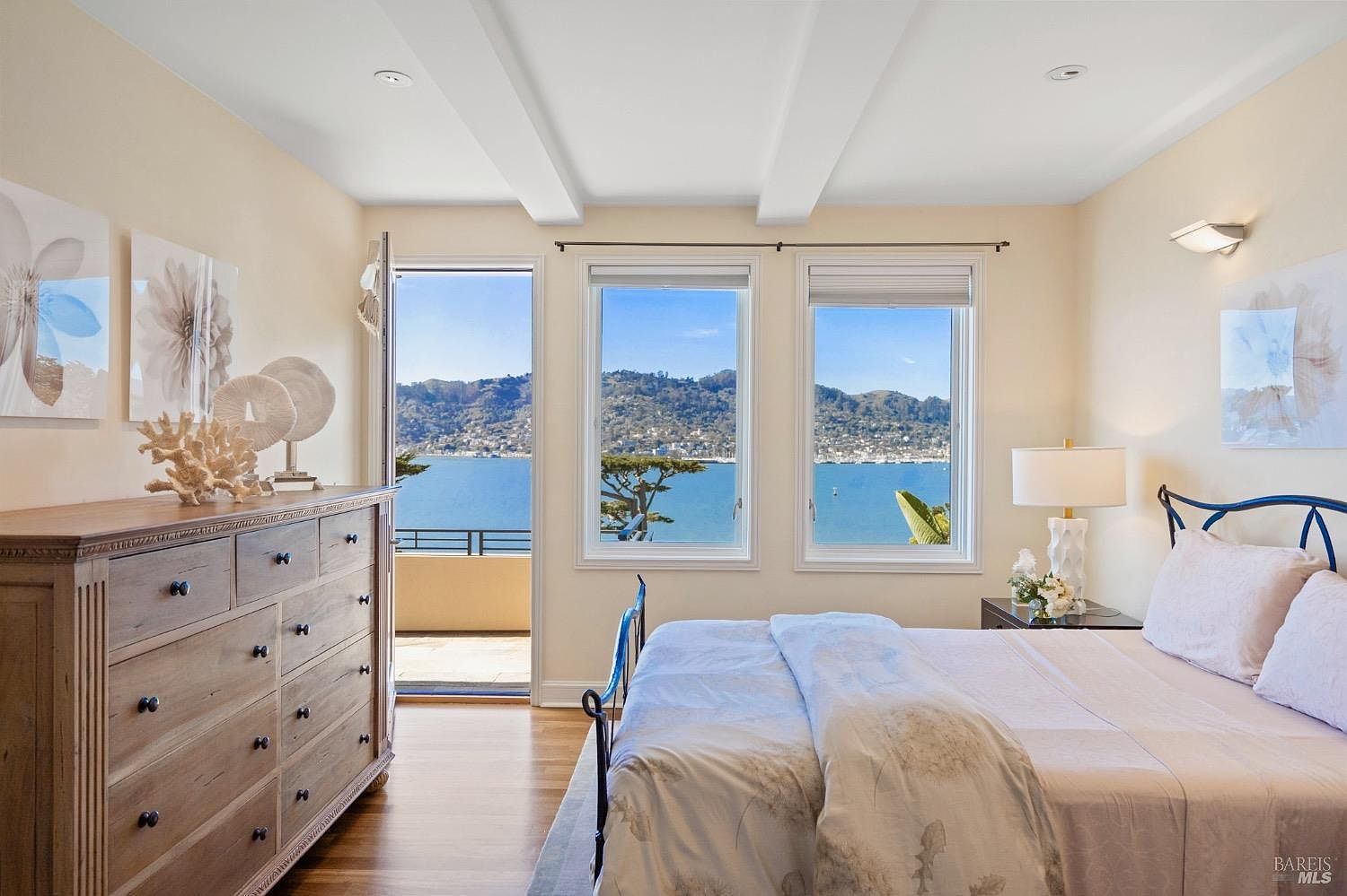 This bright and airy primary bedroom features a light wood dresser, a comfortable bed with neutral bedding, and a bedside lamp. The room is highlighted by a glass door and two large windows that provide a stunning, unobstructed view of the bay and distant hills. The space is decorated with coastal-inspired accents, creating a serene and inviting atmosphere.