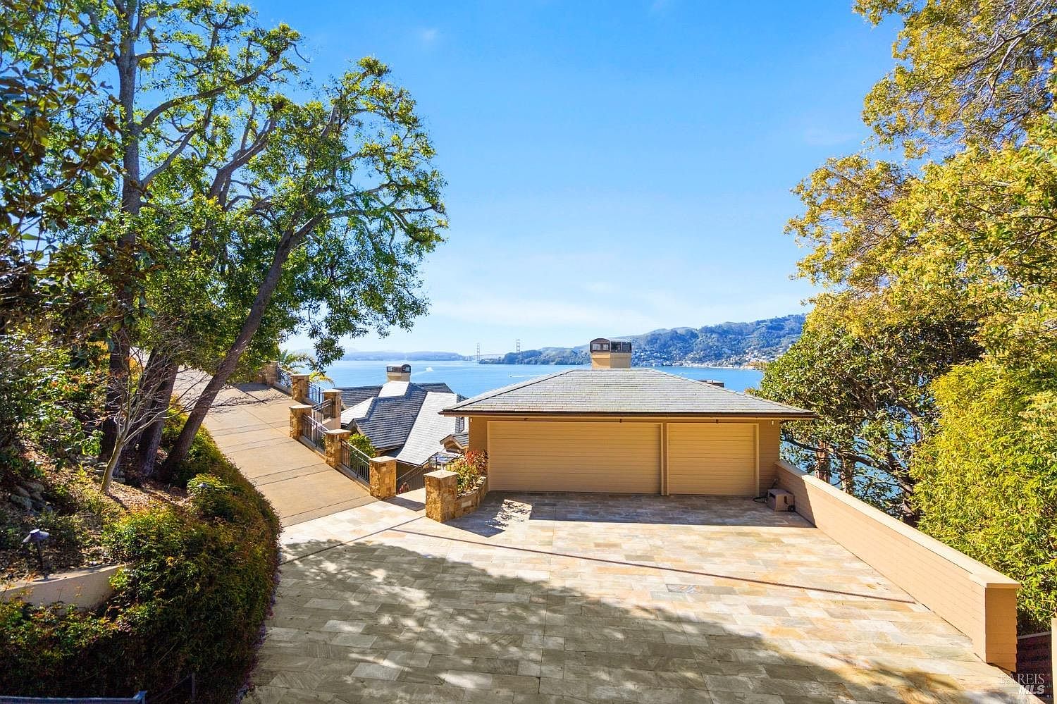 This elevated exterior view showcases a two-car garage with a paved driveway and a stone-tiled motor court. The property features a prominent view of the bay and distant hills, framed by mature trees and a stone-walled walkway leading down to the lower level. The overall impression is one of a serene, high-end coastal residence with a focus on scenic vistas.
