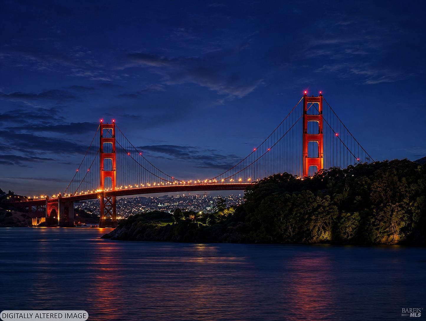 This image features a wide-angle, cinematic night view of the iconic Golden Gate Bridge illuminated against a deep blue twilight sky. The bridge's signature orange towers and suspension cables are brightly lit, casting reflections onto the dark, calm waters of the bay below. In the background, the distant city lights of San Francisco create a sparkling, atmospheric backdrop, emphasizing the property's proximity to major landmarks.