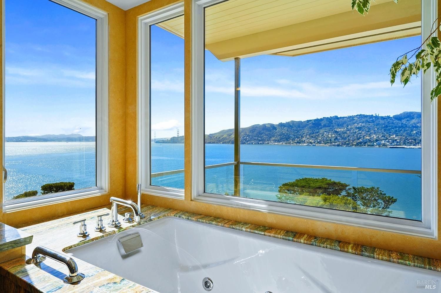 This primary bathroom features a deep soaking tub positioned directly beneath large windows that offer a panoramic view of the bay and distant hills. The tub is surrounded by a stone deck with a polished finish, complemented by modern chrome fixtures. The warm, golden-toned walls create a serene and inviting atmosphere, emphasizing the connection between the indoor space and the scenic outdoor landscape.