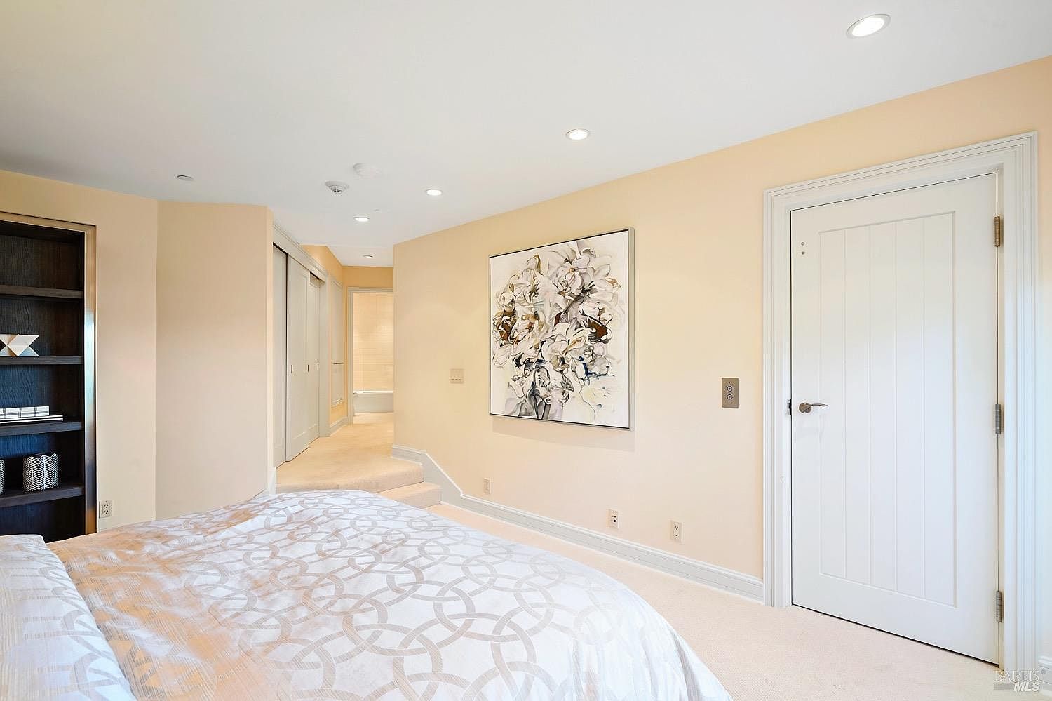 This primary bedroom features a neutral color palette with soft beige walls and plush carpeting, creating a serene and inviting atmosphere. A large, abstract floral artwork serves as a focal point on the wall, while a built-in shelving unit on the left provides elegant storage. The room includes a white paneled door and a small step-up area leading to an adjacent space, offering a sense of depth and architectural interest.