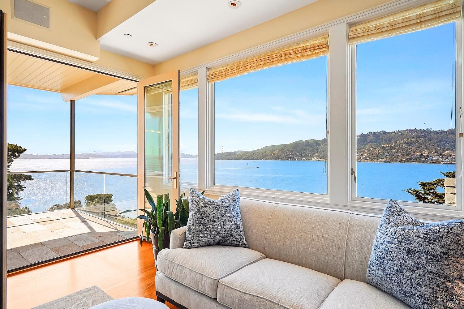 This bright living room features a neutral-toned sofa adorned with patterned throw pillows, positioned to take advantage of the expansive water views. Large windows and a glass door provide a seamless connection to an outdoor deck, flooding the space with natural light. The room is finished with warm-toned wood flooring and a potted plant, creating a serene and inviting atmosphere overlooking the bay.