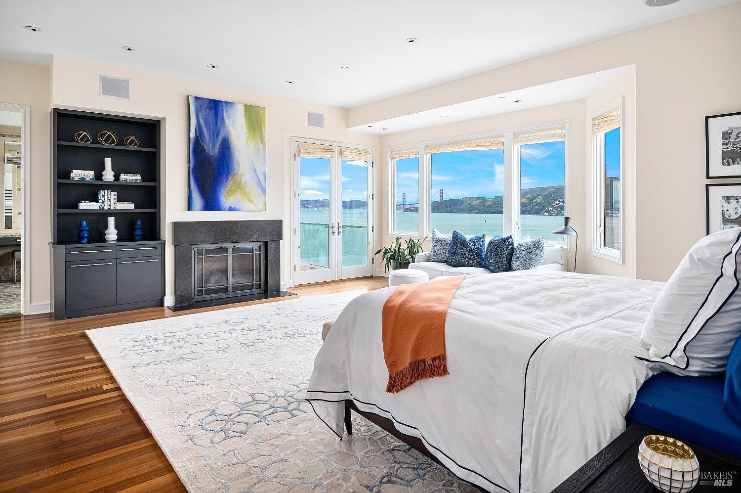 This spacious primary bedroom features a large bed with crisp white linens and a warm orange throw, positioned to face a stunning panoramic view of the bay and the Golden Gate Bridge through expansive windows. To the left, a dark-toned built-in shelving unit flanks a stone-clad fireplace, creating a sophisticated focal point against the light-colored walls. The room is anchored by a large, patterned area rug over warm-toned wood flooring, offering a serene and luxurious atmosphere.