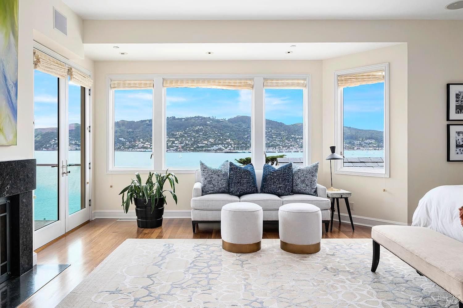 This bright and airy primary bedroom features a neutral color palette and a prominent seating area positioned before a wall of large windows that frame a scenic waterfront view. The room includes a dark stone fireplace surround on the left, a light-toned sofa with blue accent pillows, and two matching ottomans on a patterned area rug. The space is finished with warm wood flooring and minimalist decor, creating a serene and sophisticated atmosphere.