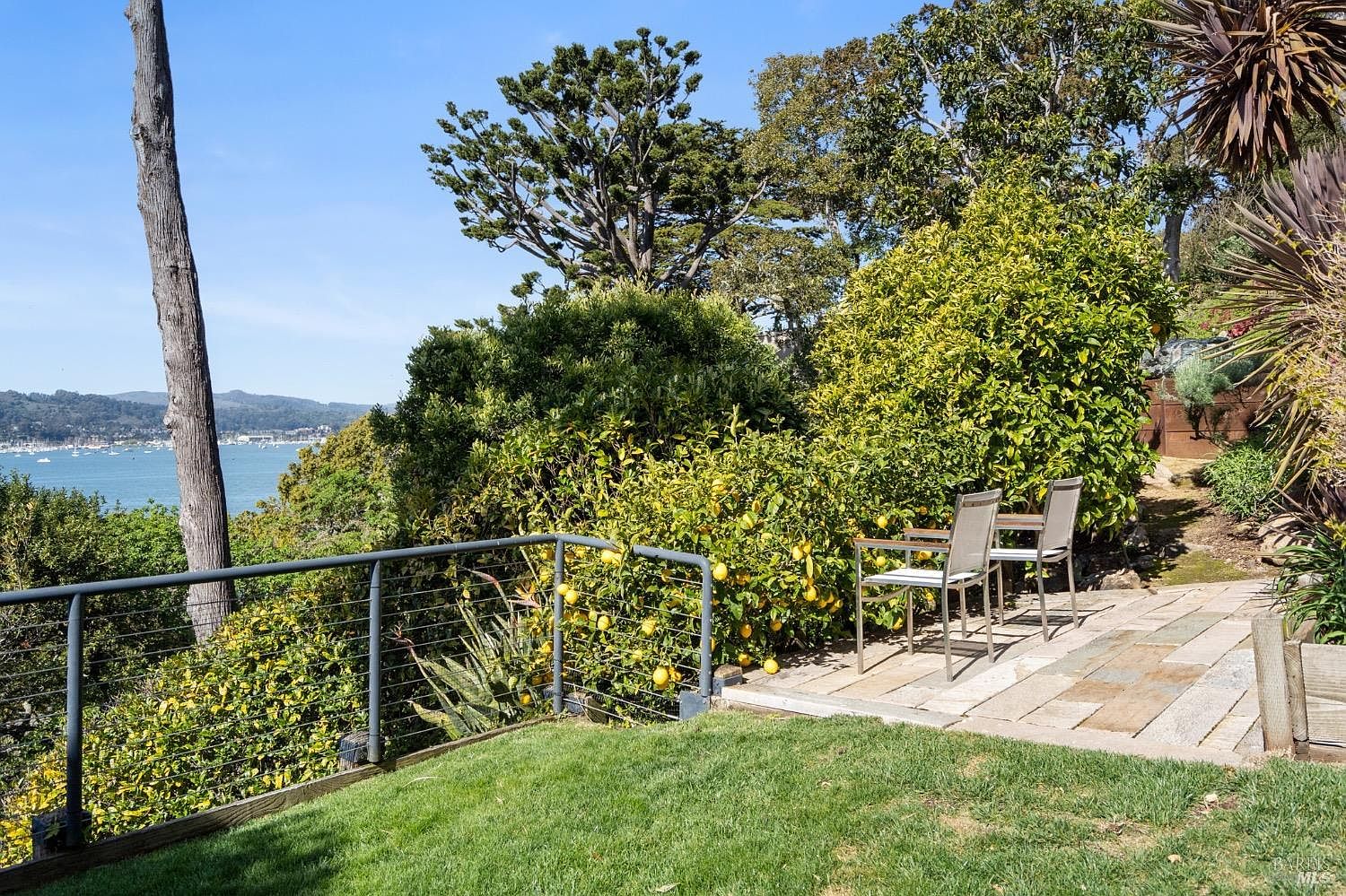 This serene outdoor space features a stone-paved patio area furnished with two chairs, nestled beside a lush lemon tree laden with fruit. A metal railing overlooks a grassy lawn and provides a stunning, elevated view of a bay filled with boats and distant hills. The setting is peaceful and private, offering an ideal spot for enjoying the coastal scenery.