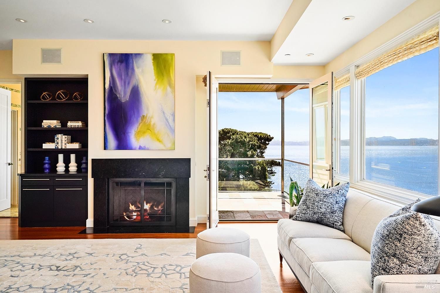 This bright and airy living room features a contemporary aesthetic with a neutral color palette, highlighted by a bold, abstract painting above a black stone fireplace. The space is anchored by a light-toned sofa and two matching ottomans, all set against a patterned area rug and warm wood flooring. Large glass doors open directly onto a balcony, framing a stunning, expansive view of the ocean and distant hills.