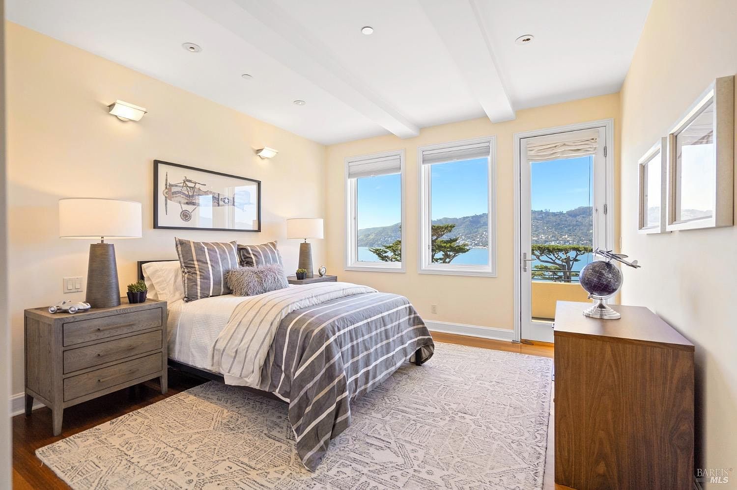 This bright and airy bedroom features a queen-sized bed dressed in striped linens, flanked by two matching wooden nightstands with modern table lamps. The room is accented by a patterned area rug, a framed airplane print on the wall, and a dresser topped with a decorative globe. Large windows and a glass-paned door provide abundant natural light and a scenic view of the water and hills beyond.