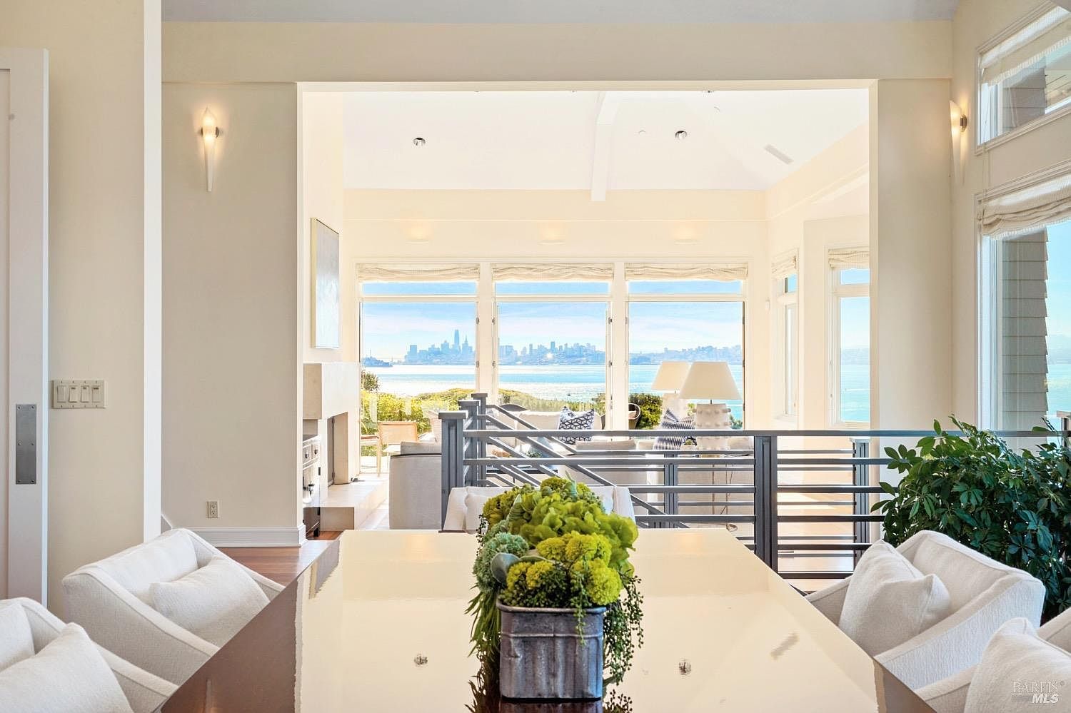 This bright dining area features a sleek, reflective table topped with a lush green centerpiece, surrounded by comfortable white upholstered chairs. The space opens up to a living area with a metal railing, leading to expansive windows that frame a stunning panoramic view of the bay and city skyline. The neutral color palette and open-concept design create an airy, sophisticated atmosphere perfect for entertaining.