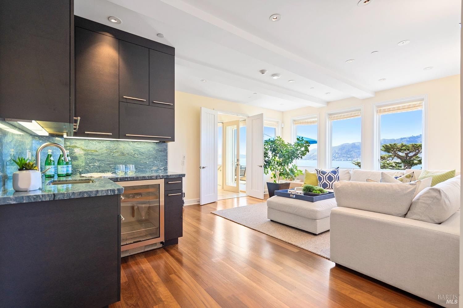 This bright and open-concept living space features a modern wet bar area with dark cabinetry and a stone backsplash, seamlessly transitioning into a comfortable seating area. Large windows provide expansive views of the water and hills, while double doors open to an outdoor space, enhancing the indoor-outdoor flow. The room is finished with warm wood flooring and a neutral color palette, creating an inviting and airy atmosphere.
