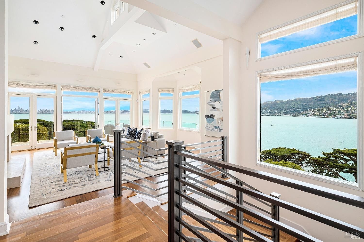 This bright, open-concept living area features a vaulted ceiling and expansive windows that frame panoramic water and city skyline views. The space is furnished with light-toned seating arranged on a patterned area rug, centered around a fireplace with a tall surround. A dark metal railing separates the living area from the foreground staircase, creating a sophisticated and airy atmosphere.