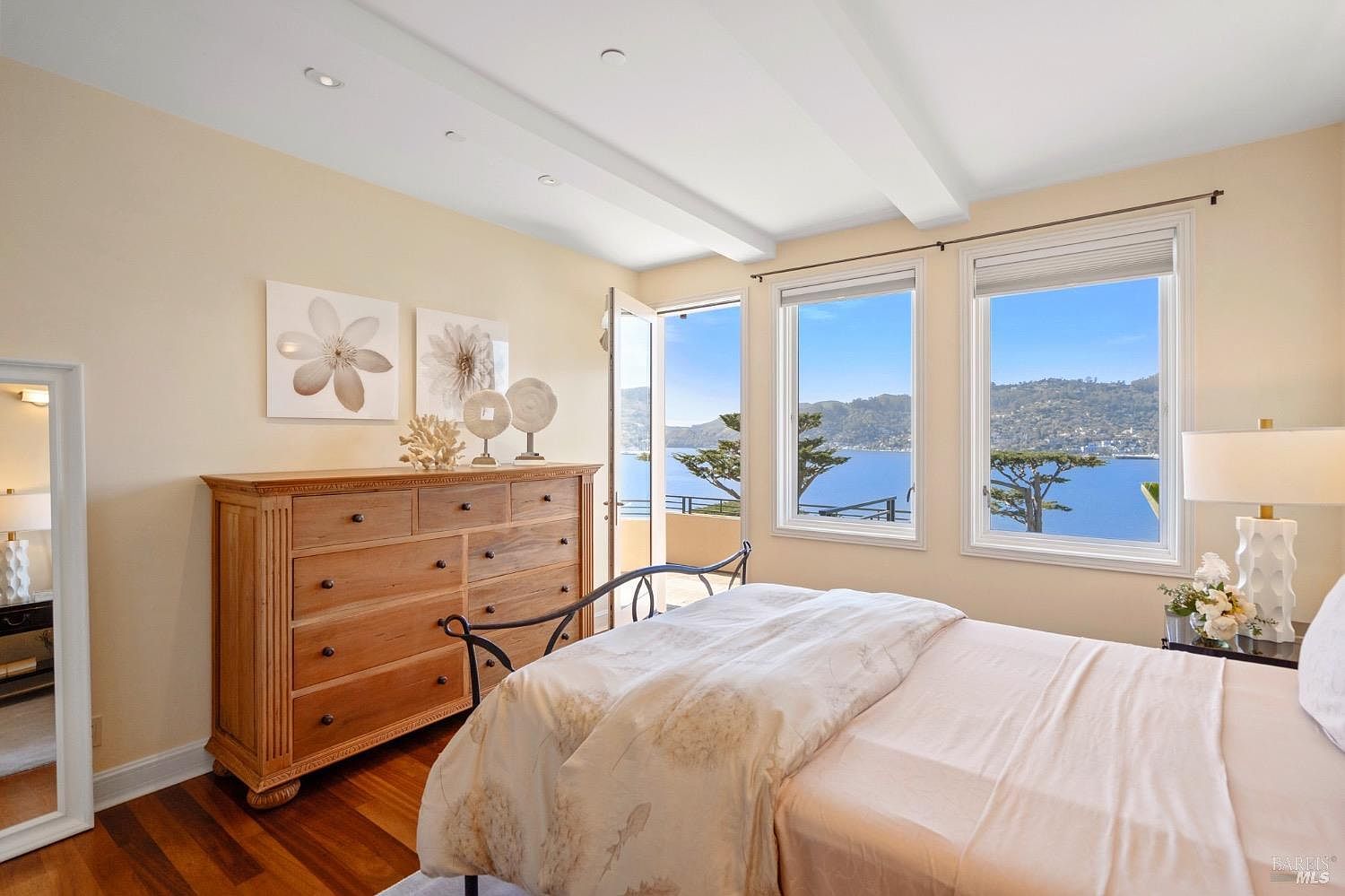 This bright and airy bedroom features a light wood dresser, a bed with neutral bedding, and a full-length mirror. Large windows provide an expansive, panoramic view of the water and distant hills, creating a serene and tranquil atmosphere. The room is decorated with minimalist floral wall art and elegant table lamps, emphasizing a clean and sophisticated aesthetic.