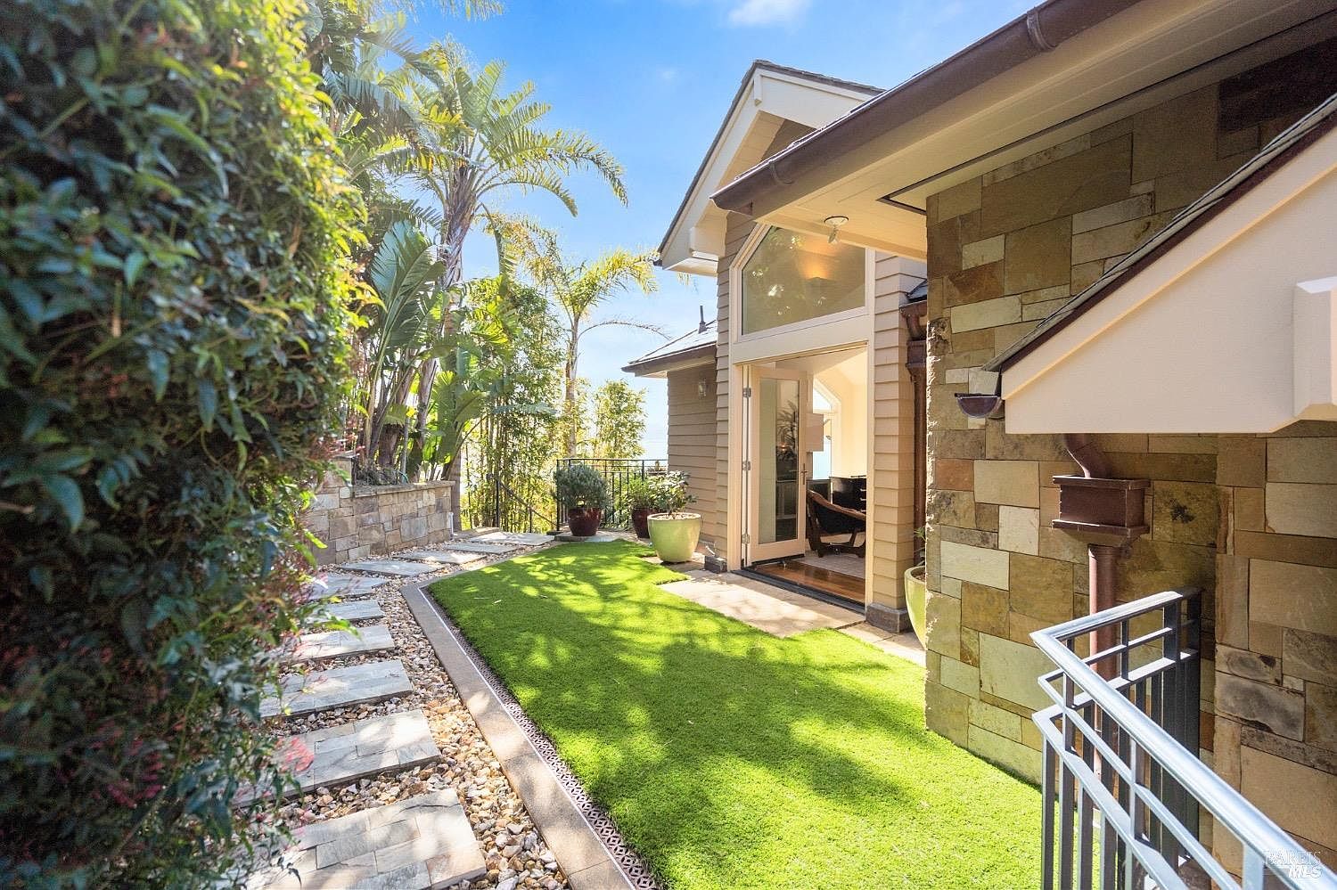 This inviting outdoor space features a lush green lawn bordered by a stone-paved walkway and a stone-clad exterior wall. A set of glass doors opens from the home onto the yard, creating a seamless transition between indoor and outdoor living areas. The scene is framed by mature palm trees and lush greenery, offering a serene and private atmosphere.