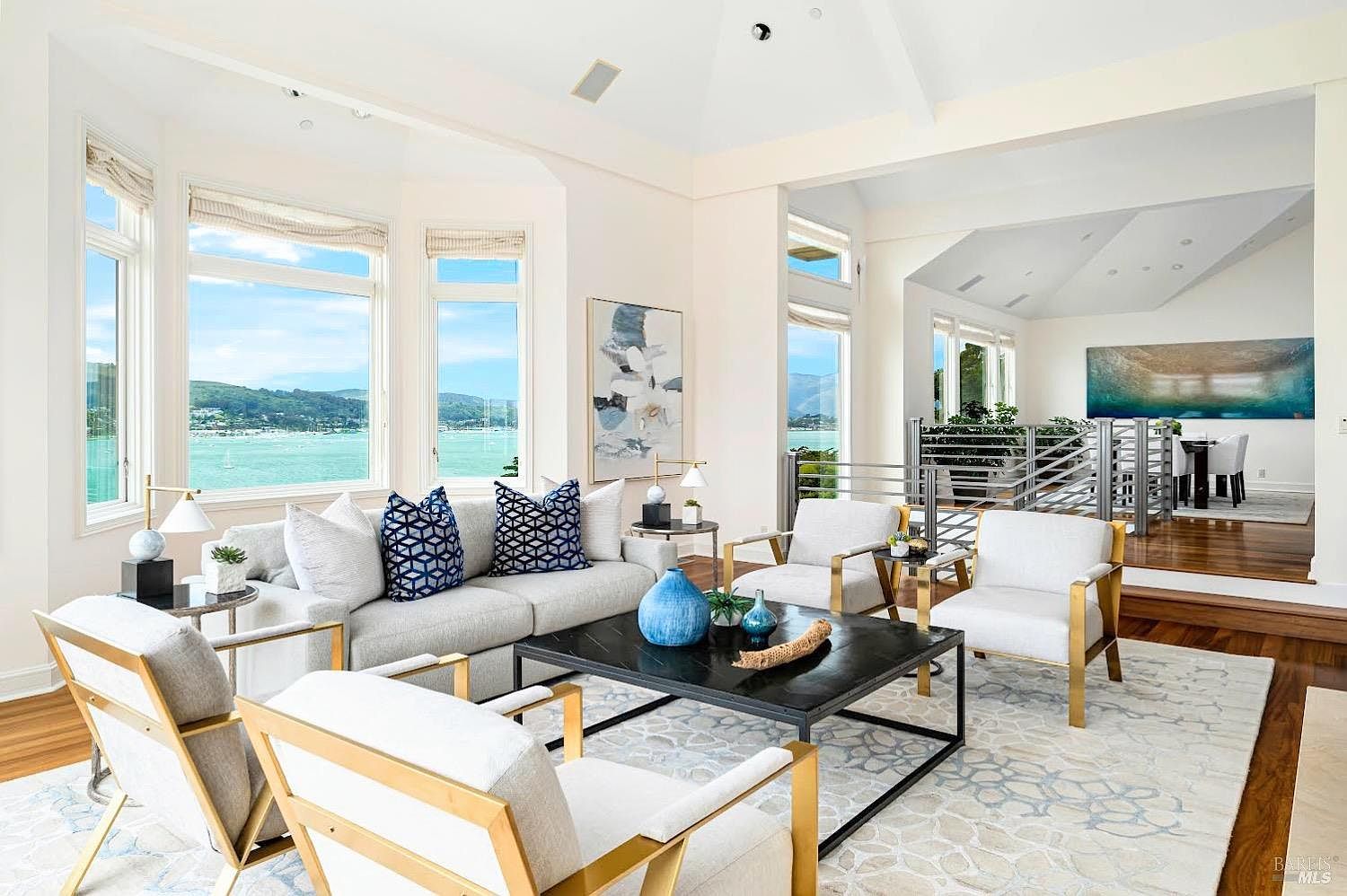 This bright and airy living room features a sophisticated, neutral color palette with pops of blue in the accent pillows and decor. The space is anchored by a large, dark-toned coffee table and surrounded by comfortable seating, all positioned to take advantage of the expansive windows overlooking the water. A raised dining area is visible in the background, connected by a sleek metal railing, creating an open and cohesive floor plan.