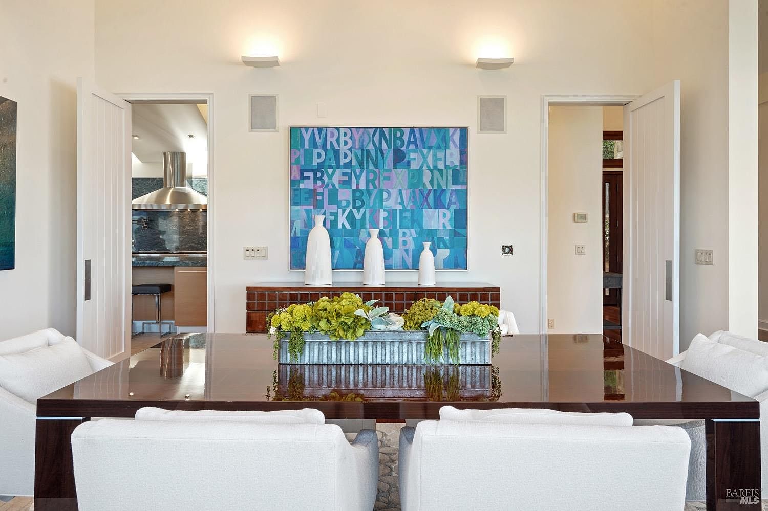 This elegant dining room features a large, dark-toned table centered in the frame, topped with a long, rectangular planter filled with lush green foliage. Behind the table, a sideboard holds three minimalist white vases beneath a vibrant, abstract letter-art piece mounted on the wall. The room is framed by two open doorways leading to adjacent spaces, with soft, warm lighting emanating from wall-mounted fixtures above the artwork.