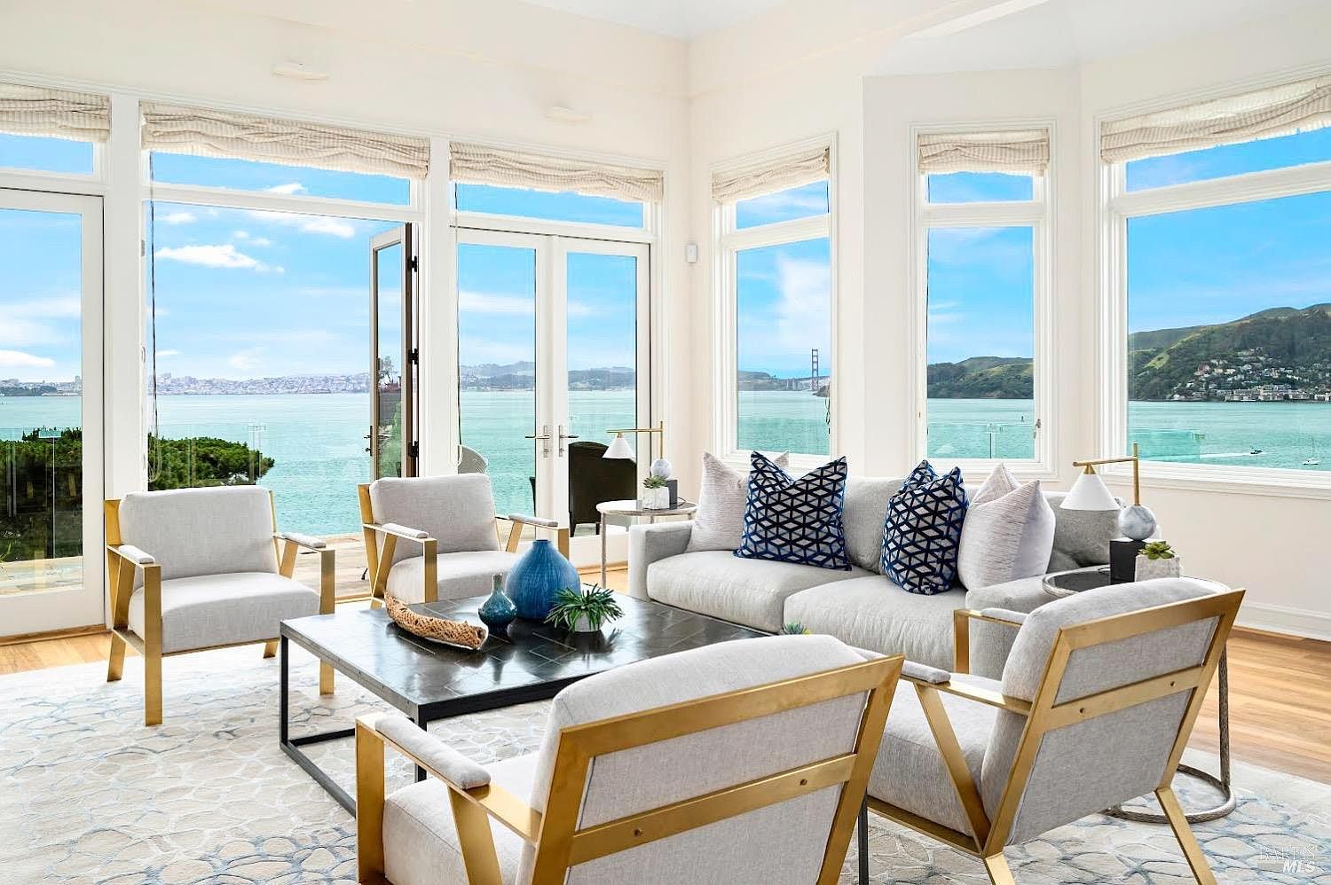 This bright and airy living room features a sophisticated seating arrangement centered around a dark coffee table, complemented by light-toned armchairs and a sofa with patterned accent pillows. The space is defined by expansive windows that offer panoramic views of the bay and distant hills, creating a seamless connection between the interior and the waterfront scenery. The room's neutral color palette and metallic-framed furniture contribute to a refined, contemporary aesthetic.