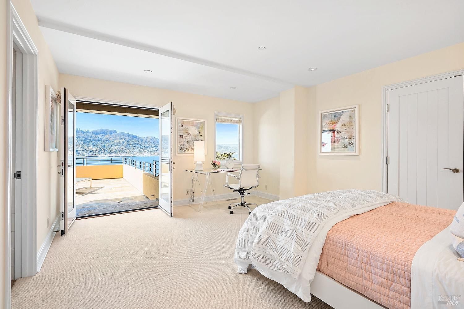This bright and airy bedroom features a neutral color palette, plush carpeting, and a bed dressed in light-toned bedding. The room opens directly onto a spacious balcony through double glass doors, offering a stunning, unobstructed view of the water and distant hills. A sleek glass-topped desk is positioned near the window, creating a functional workspace that takes advantage of the natural light and scenic backdrop.