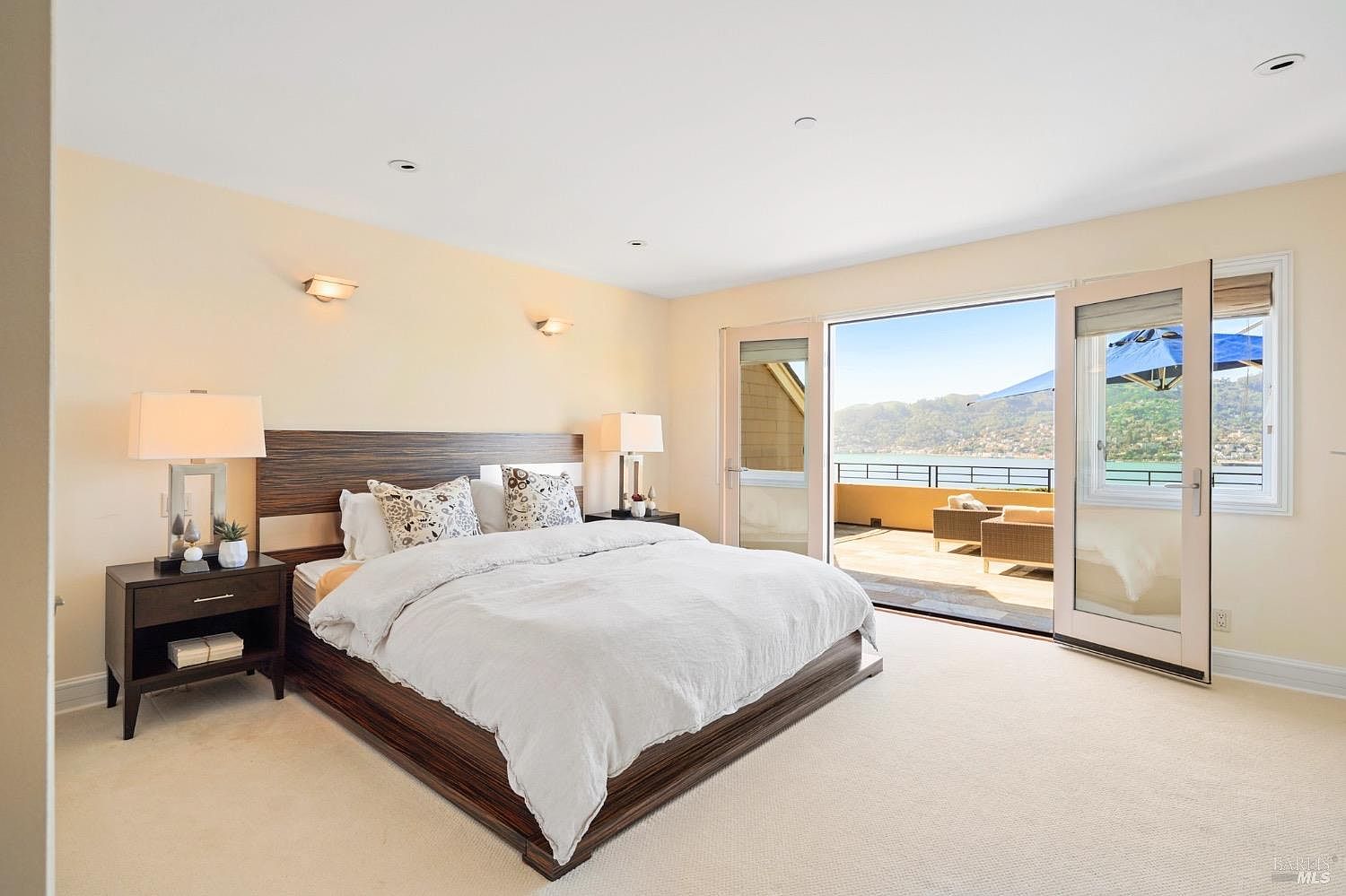 This spacious primary bedroom features a modern platform bed with a dark wood headboard and neutral bedding, flanked by matching nightstands and wall-mounted lamps. Large glass doors open directly onto a private balcony, providing an expansive, unobstructed view of the water and surrounding hills. The room is finished with light-colored walls and carpeting, creating a bright and serene atmosphere.
